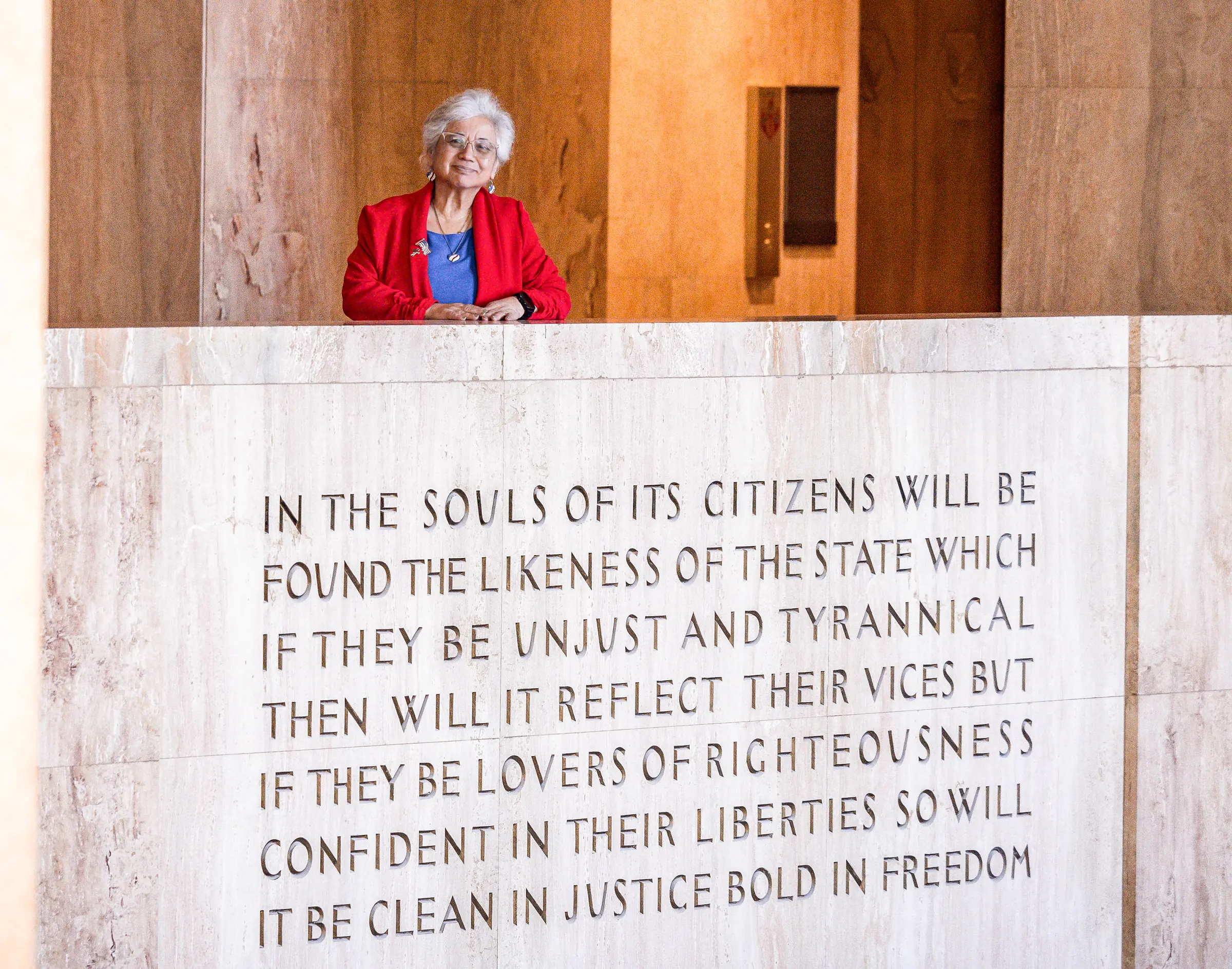 Anna Munson in a red jacket inside the Oregon State Capitol, standing behind a marble wall engraved with the Capitol’s inscription about citizens and the state
