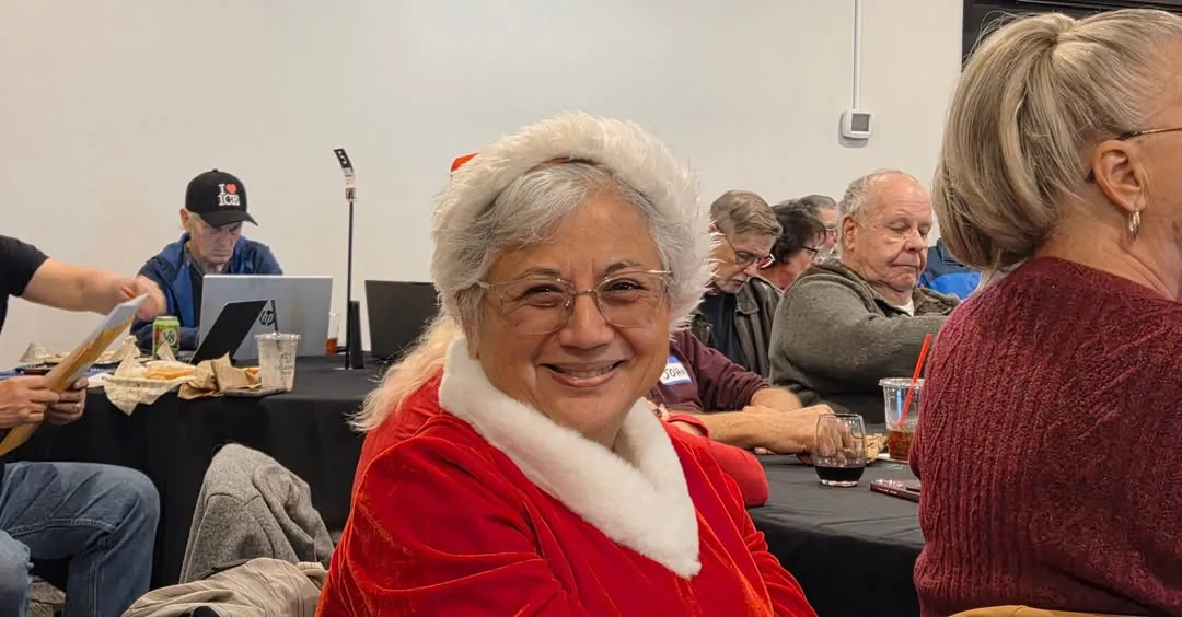 Anna Munson smiling in a red velvet Santa suit with white fur trim and a white fur headband, seated at a table at a community gathering